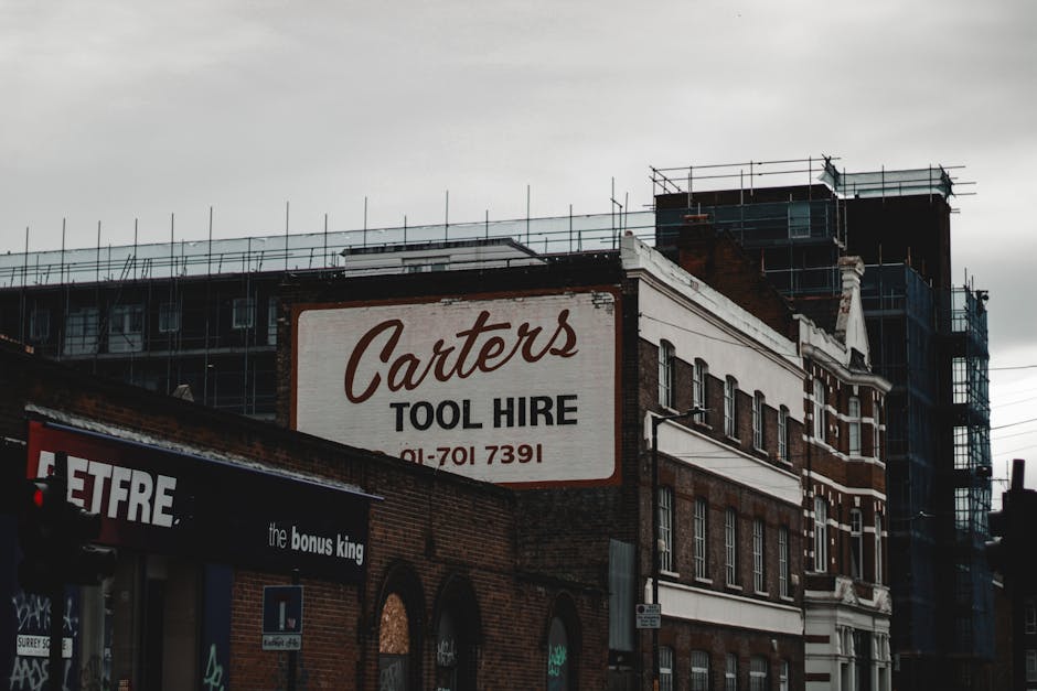 A multi-story brick building with scaffolding on its upper floors, indicating ongoing construction or refurbishment. Attached to the building is a large rectangular sign for 'Carter's Tool Hire' displayed prominently on the facade, featuring bold black lettering and a phone number. The lower part of the building houses a storefront with a black awning labeled 'the bonus king' and some graffiti on the wall beneath it. The scene is set outdoors on a cloudy day, with the building facing a street or pavement, surrounded by urban elements signifying the area near Bermondsey Station and Maltby Market, suitable for house removals and furniture transport activities by Man and a Van Bermondsey.